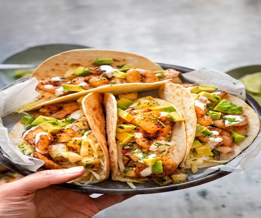 A hand holding a plate of shrimp tacos with avocado and cabbage slaw