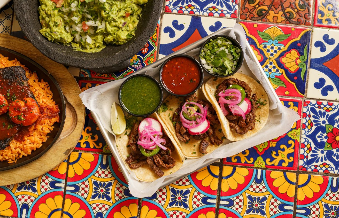 A platter of grilled beef tacos with salsas, guacamole, and shrimp on a colorful tiled table